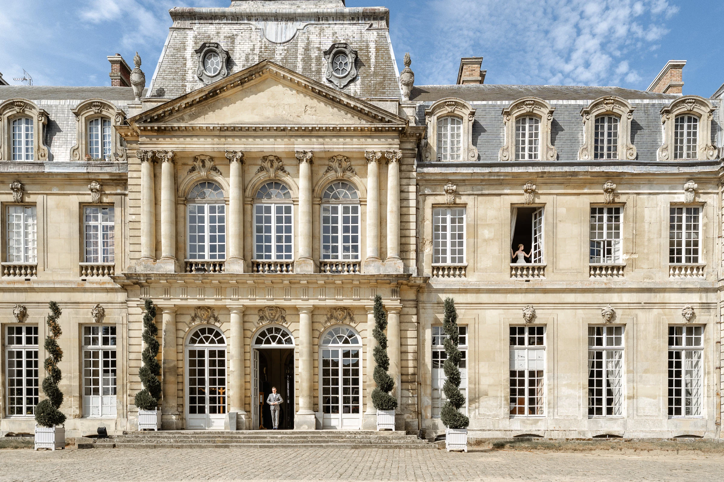 La façade du château de Champlâtreux, le marié sur le perron et la mariée à la fenêtre