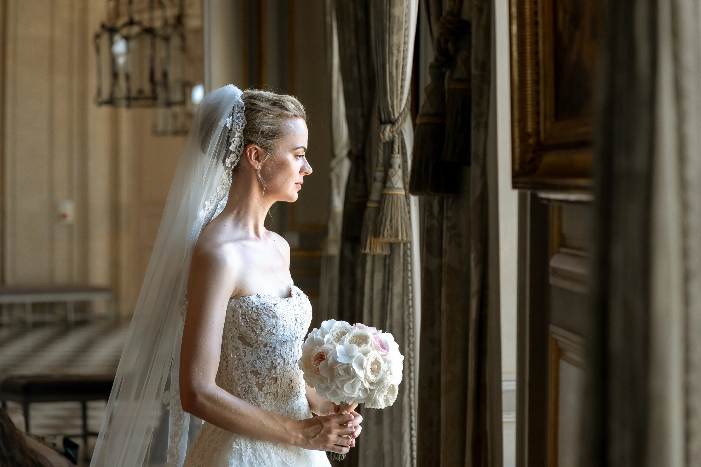 The bride in profile, bouquet in hand, between the drawing-room curtains, château de Champlâtreux