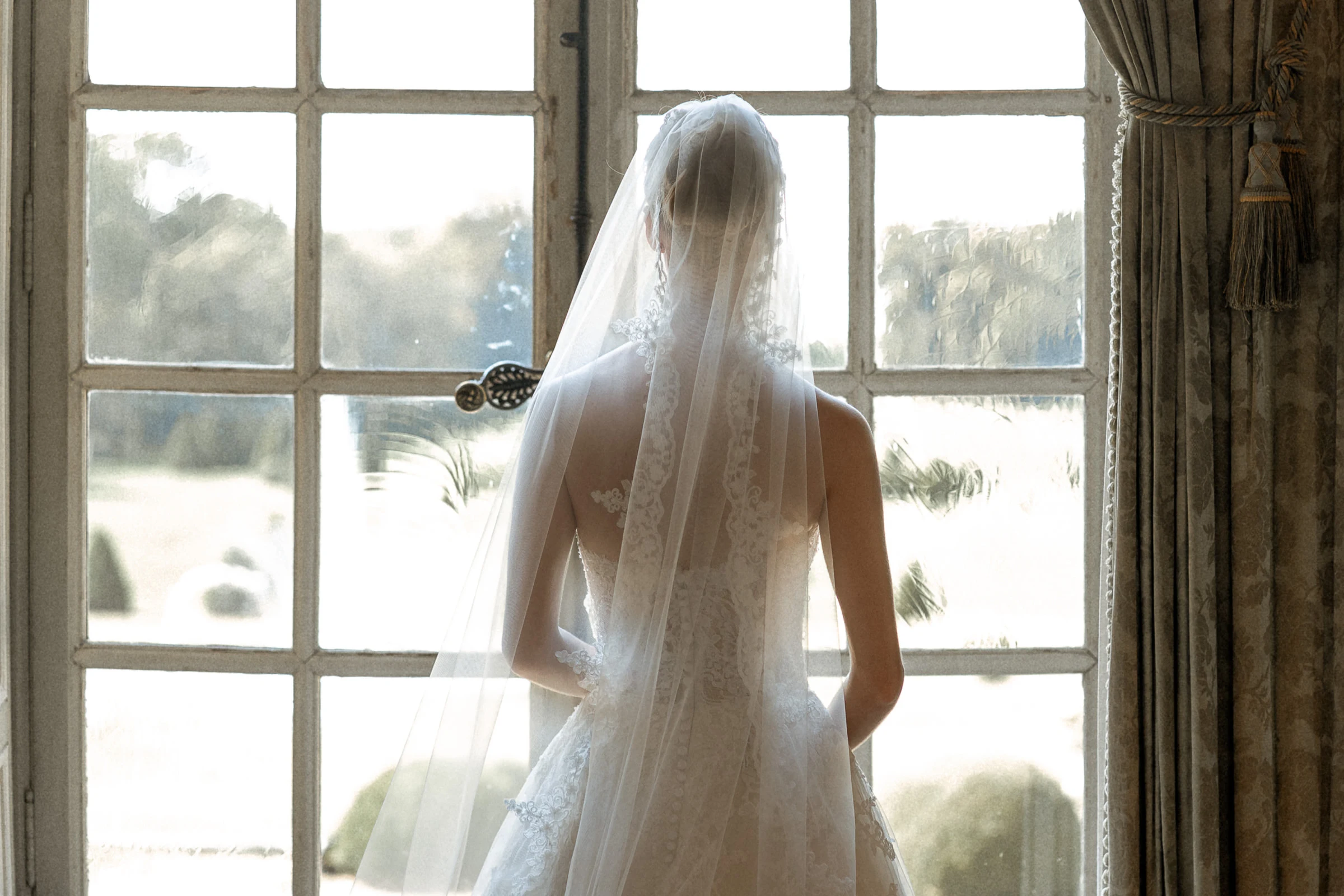 La mariée de dos sous son voile devant la grande fenêtre ouverte sur le parc, château de Champlâtreux